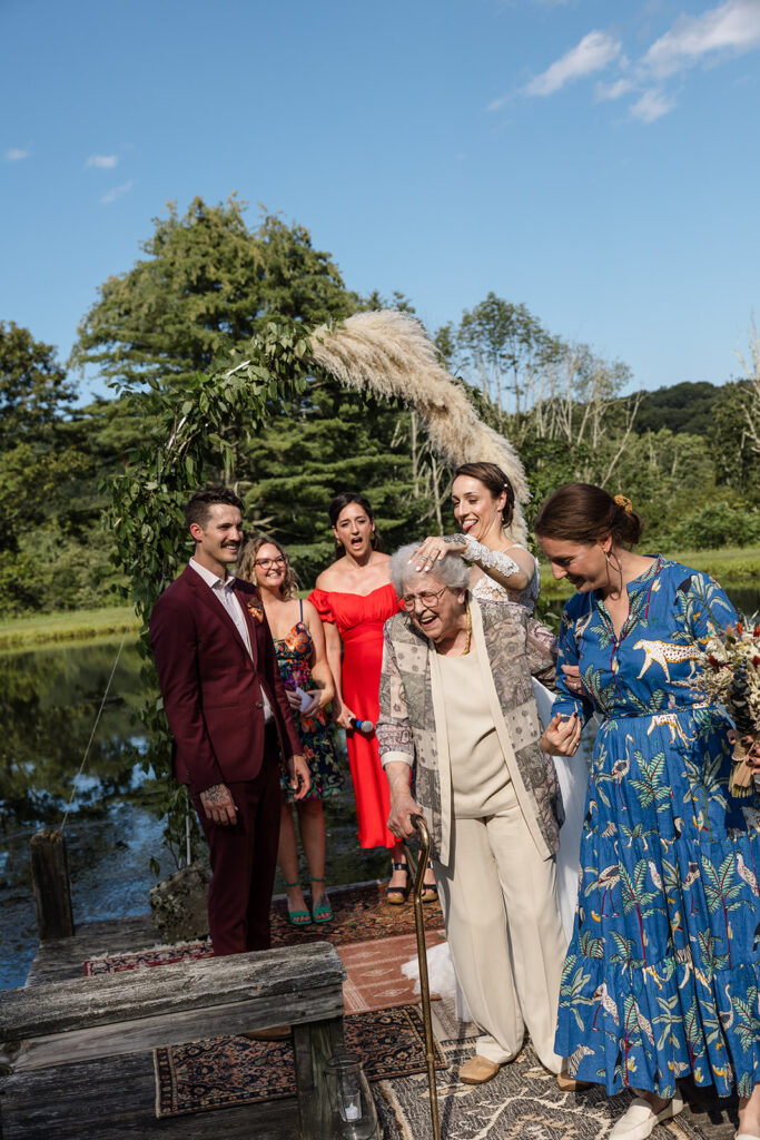 A group of wedding guests, including the bride and groom, gather around an elderly woman as she walks away from the wedding ceremony, joyfully celebrating the moment outdoors by a pond.