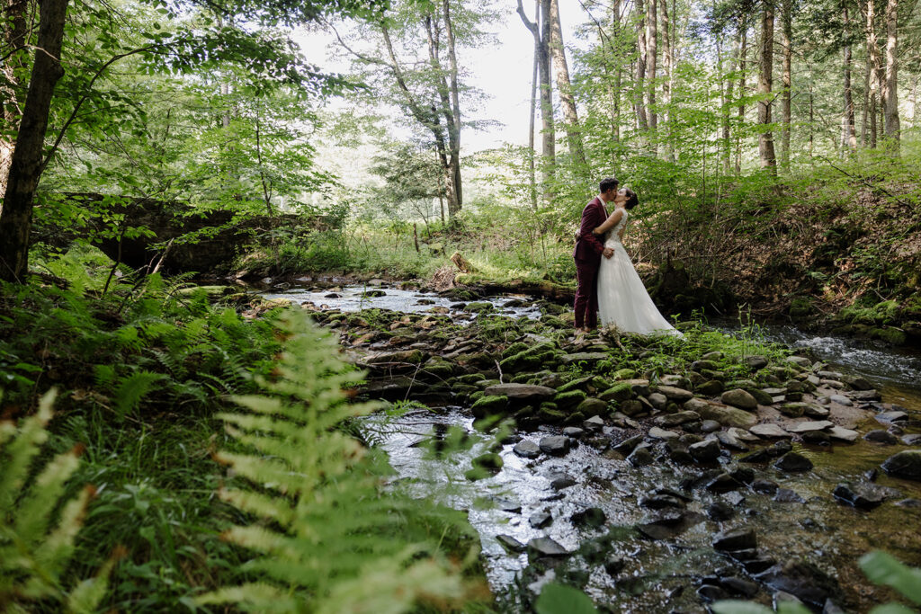 A newlywed couple shares a kiss while standing on a moss-covered, rocky streambed surrounded by a dense forest, with dappled sunlight filtering through the trees.