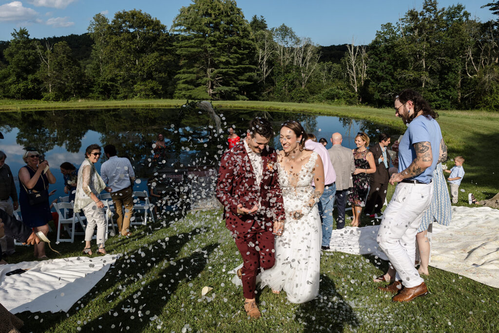 The bride and groom walk hand-in-hand down the aisle, surrounded by guests who shower them with confetti, creating a joyful and festive atmosphere by a scenic pond.