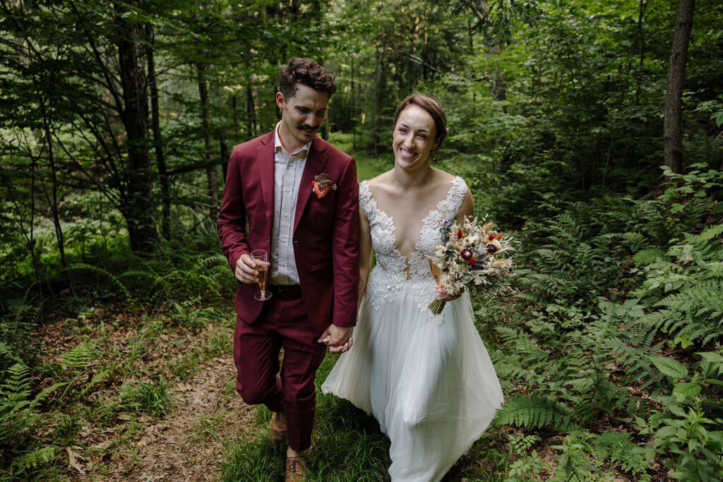 A couple strolls hand in hand through a forest. The groom is dressed in a burgundy suit and carries a glass of champagne, while the bride wears a white lace wedding dress with a deep V neckline, holding a dried floral bouquet. They both smile as they walk through the lush greenery.