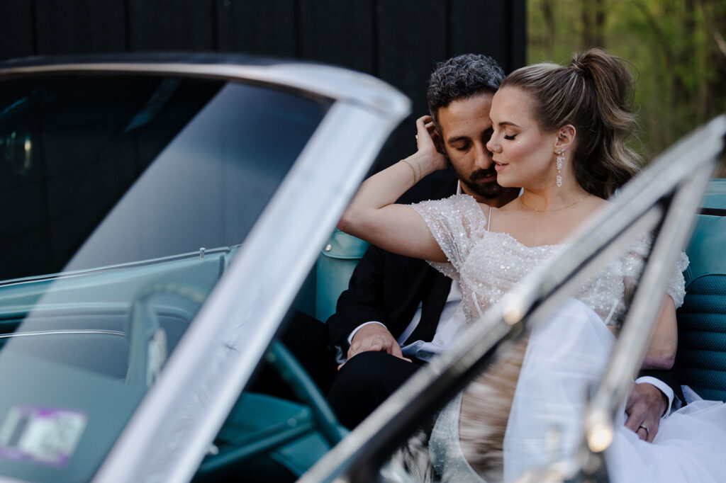 A bride and groom share an intimate moment in the front seat of a classic turquoise convertible car. The bride, in an off-shoulder gown with sparkling beadwork, rests her head on the groom's shoulder, while he gazes at her lovingly. The couple's relaxed and tender pose captures their connection.