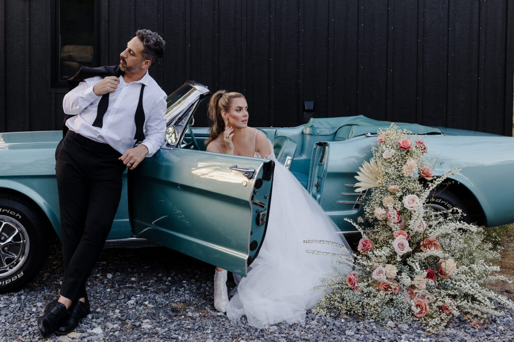 A groom leans casually against the open door of a turquoise classic car, holding his jacket over his shoulder, while the bride sits inside the car. The bride’s dress flows out of the car, and an arrangement of pastel flowers decorates the side, enhancing the vintage vibe of the scene.