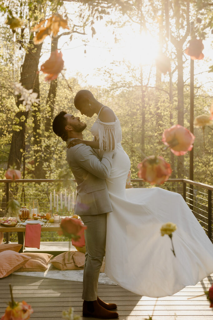 A couple enjoys a romantic moment outdoors, surrounded by hanging flowers illuminated by golden sunlight. The groom lifts the bride in his arms, both smiling and gazing into each other’s eyes. The dreamy setting with suspended blooms and warm light creates an ethereal ambiance.