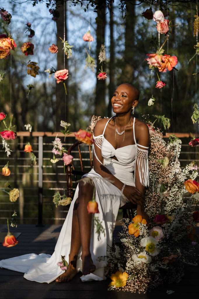 The bride sits gracefully on a chair amidst a floral installation, surrounded by hanging flowers of various colors. She wears a white dress with fringed sleeves, looking off to the side with a radiant smile. The sunlight streaming in enhances the beauty of the scene.