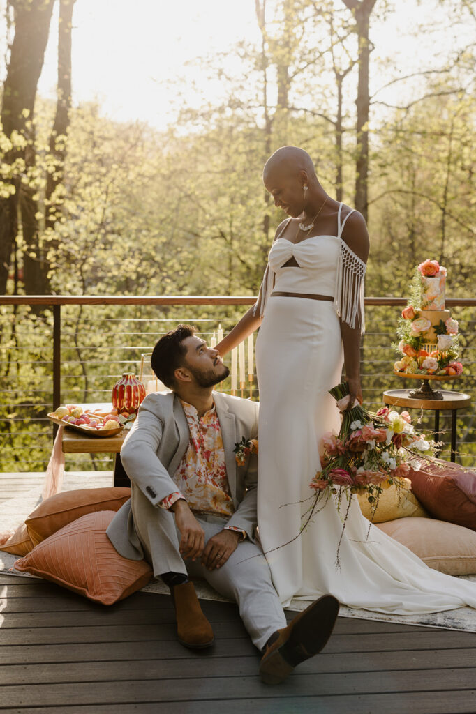 The bride stands elegantly while the groom sits at her feet, gazing up at her. She holds a bouquet of pink and white flowers, and her off-shoulder dress features intricate fringe detailing. A table with fruits and candles is visible in the background, adding to the cozy atmosphere.