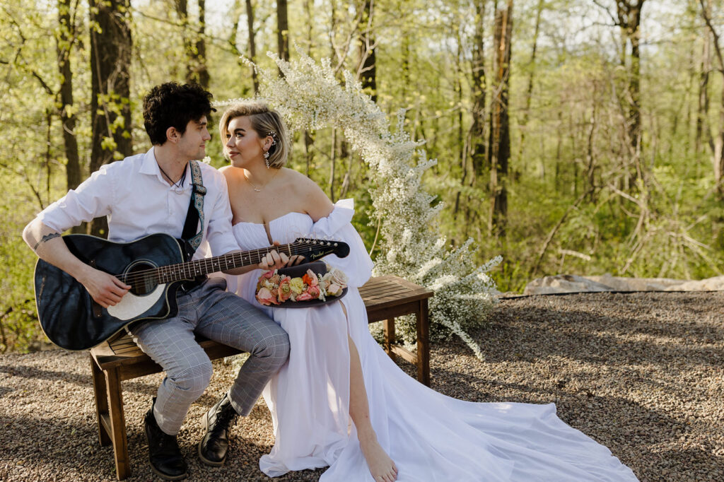 A couple sits on a bench in a wooded area, with the groom playing a guitar. The bride, in an off-shoulder gown, gazes lovingly at him, holding a small bouquet of flowers. A delicate floral arch frames the scene, adding to the romantic ambiance.