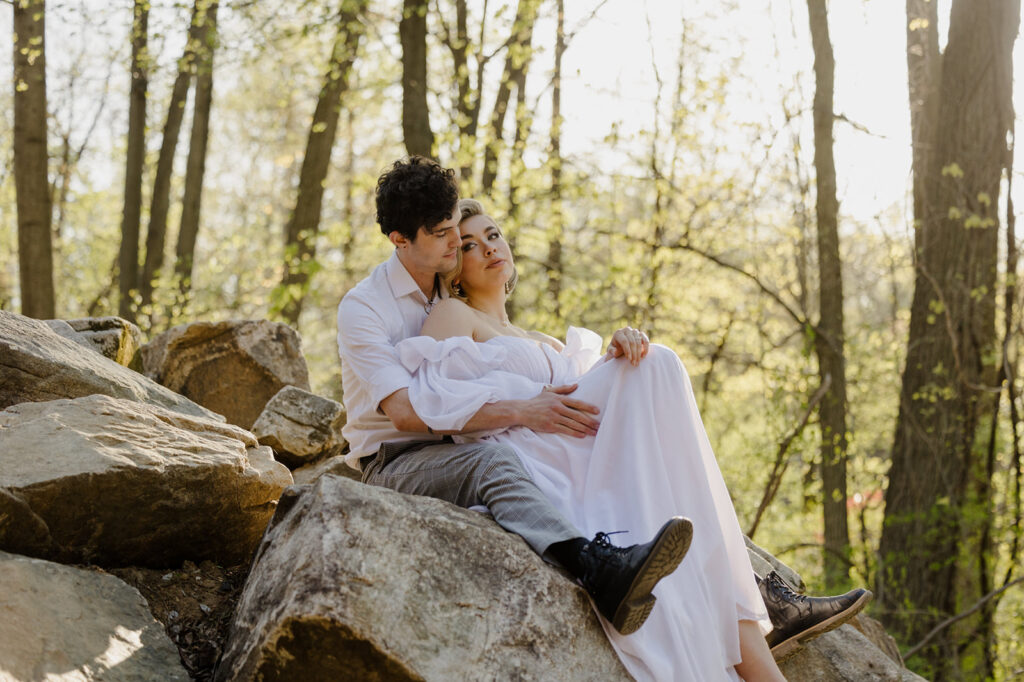 The bride and groom sit on large rocks, nestled in a forest. The bride leans back into the groom’s embrace, her white gown cascading over the rocks, while the groom, dressed casually, looks off into the distance. The sunlight filtering through the trees casts a soft glow over the couple. ​