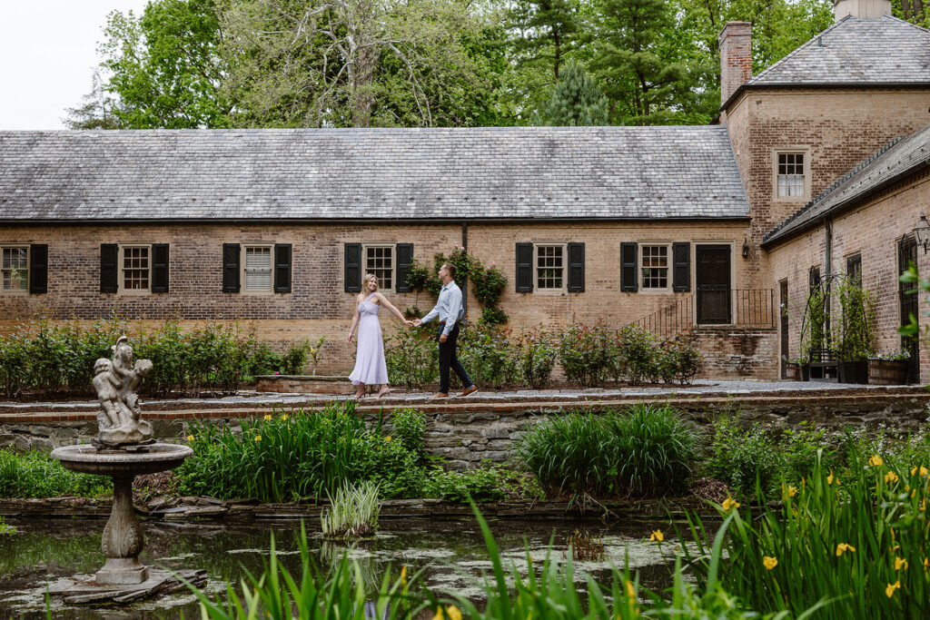 A couple walks hand in hand along the garden path of Conestoga House, surrounded by a manicured landscape of shrubs and blooming flowers. The building in the background is a classic, historic brick structure with dark shutters, enhancing the romantic and elegant atmosphere of this engagement or wedding photo.