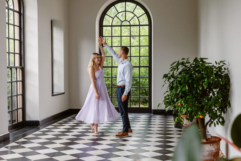 The couple dances gracefully inside a beautifully lit hall with large arched windows at Conestoga House. The bride’s lavender dress flows elegantly as her partner twirls her. The checkered black-and-white floor and a potted orange tree create a sophisticated backdrop, combining indoor charm with hints of nature.