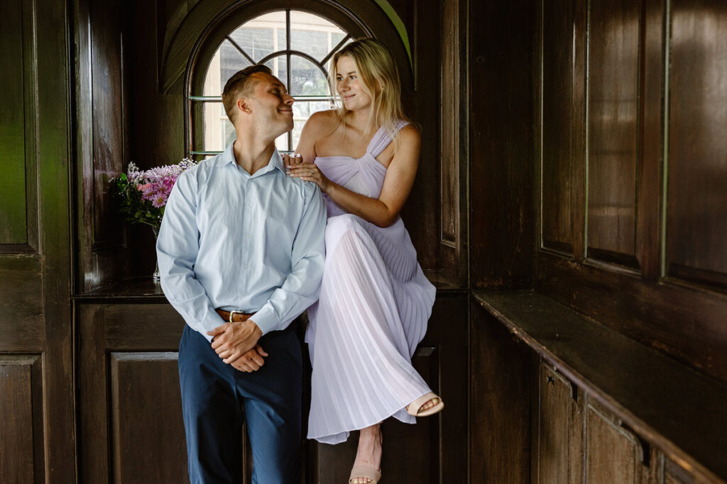 The couple shares a quiet moment together on a wooden bench surrounded by dark wooden paneling. The bride, in her soft, pleated lavender dress, sits slightly elevated while gazing down lovingly at her partner, who stands close by. The light filtering through the arched window creates a warm and romantic ambiance.