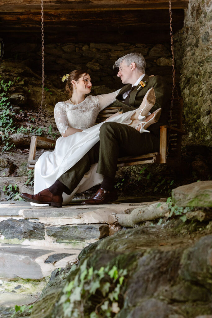 The bride and groom sit on a rustic wooden swing, surrounded by stone walls and greenery at Drumore Mill. The bride’s delicate lace dress and the groom’s formal attire contrast with the rugged textures of the stone and wood, capturing a beautiful blend of elegance and rustic charm.