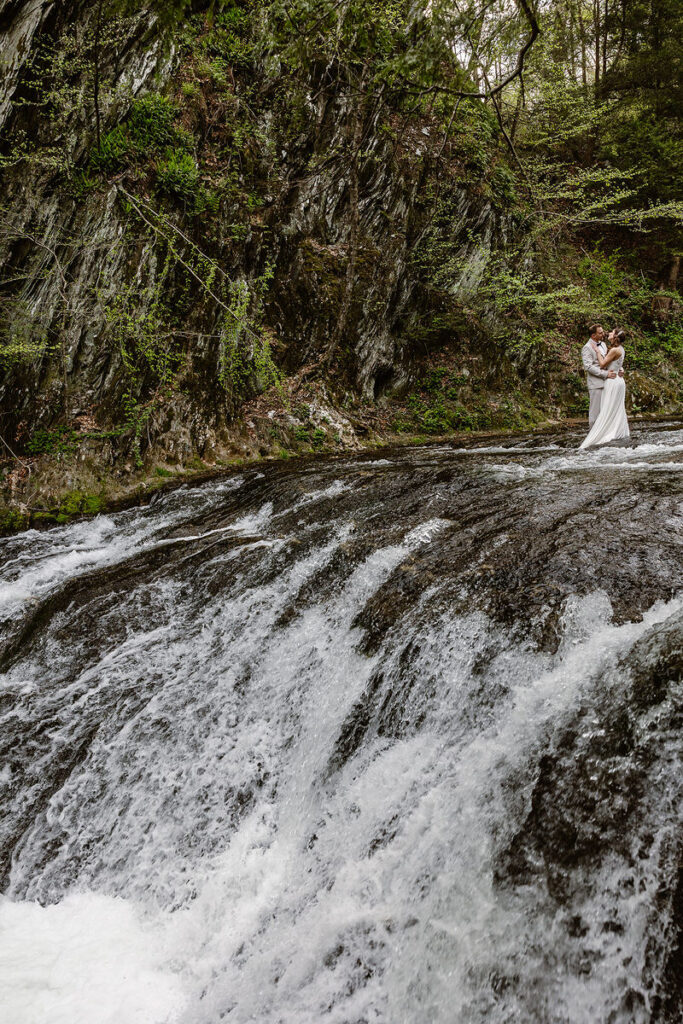 The couple is seen standing on a rock ledge above a cascading waterfall, enveloped by lush, towering cliffs. The bride’s flowing white dress catches the breeze, and the groom holds her close, creating a dramatic and adventurous feel to this wedding portrait.