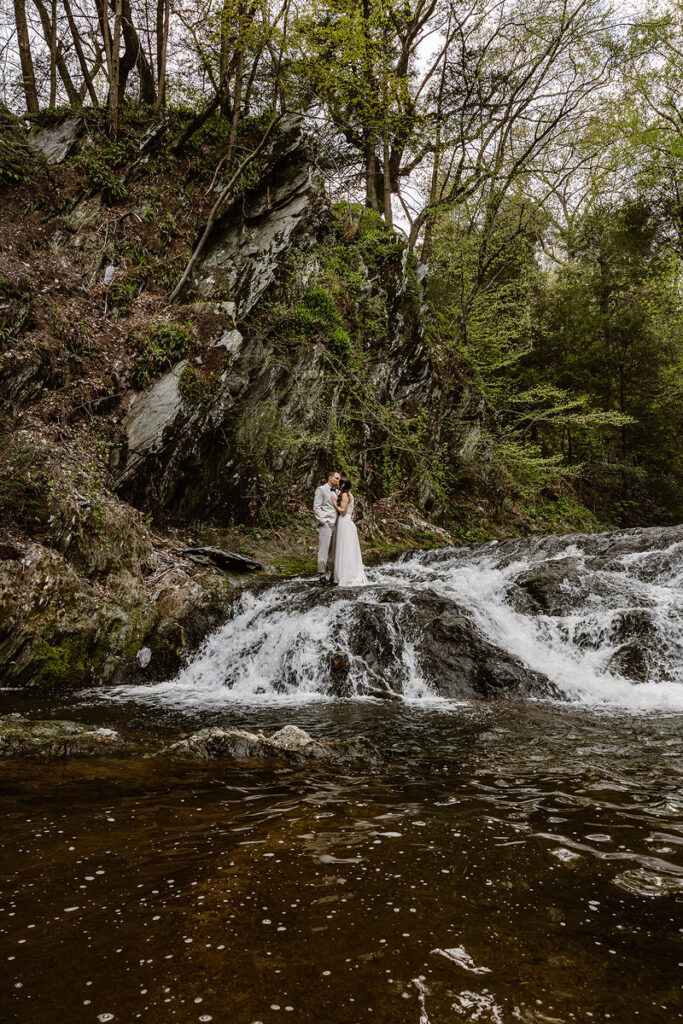 The couple stands together on a rocky surface near the base of a waterfall, surrounded by the serene sounds of rushing water. The natural beauty of the rocky terrain and dense greenery add an element of wild romance to this picturesque scene.