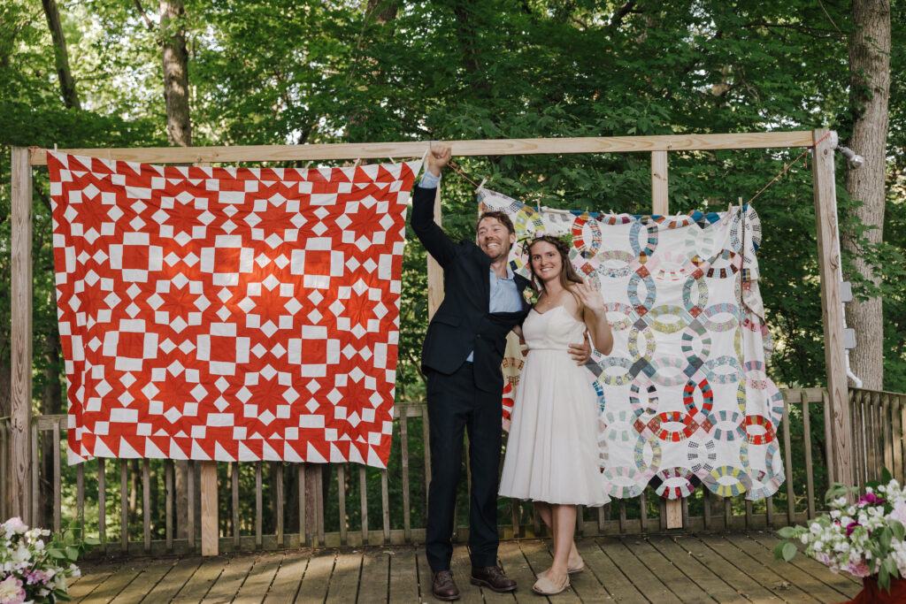 A couple standing in front of two colorful quilts hanging on a wooden frame. The groom is in a dark suit and playfully holds up the red and white quilt, while the bride, in a knee-length white dress, smiles beside him. They are outdoors, with a forest in the background.