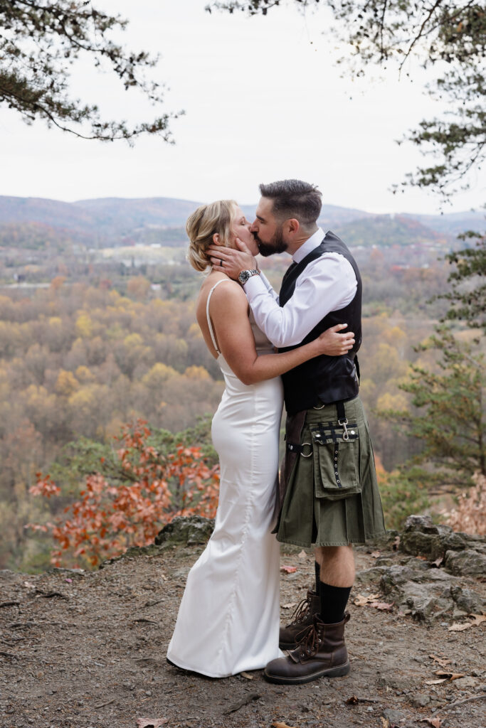 A bride and groom sharing a kiss in a scenic outdoor location with hills and trees in the background. The groom wears a vest and a kilt, while the bride is in a sleek white gown.