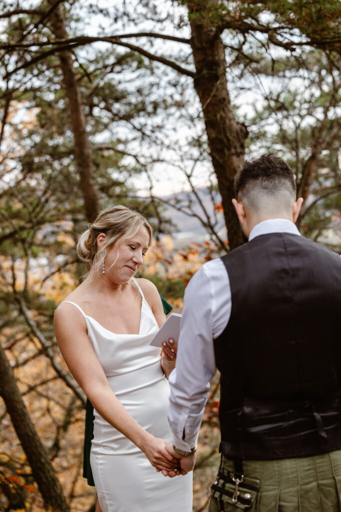 A couple exchanging vows in a forest setting. The bride wears a simple, elegant white dress and reads from a small notebook, while the groom, wearing a black vest and shirt, stands facing her, holding her hand.