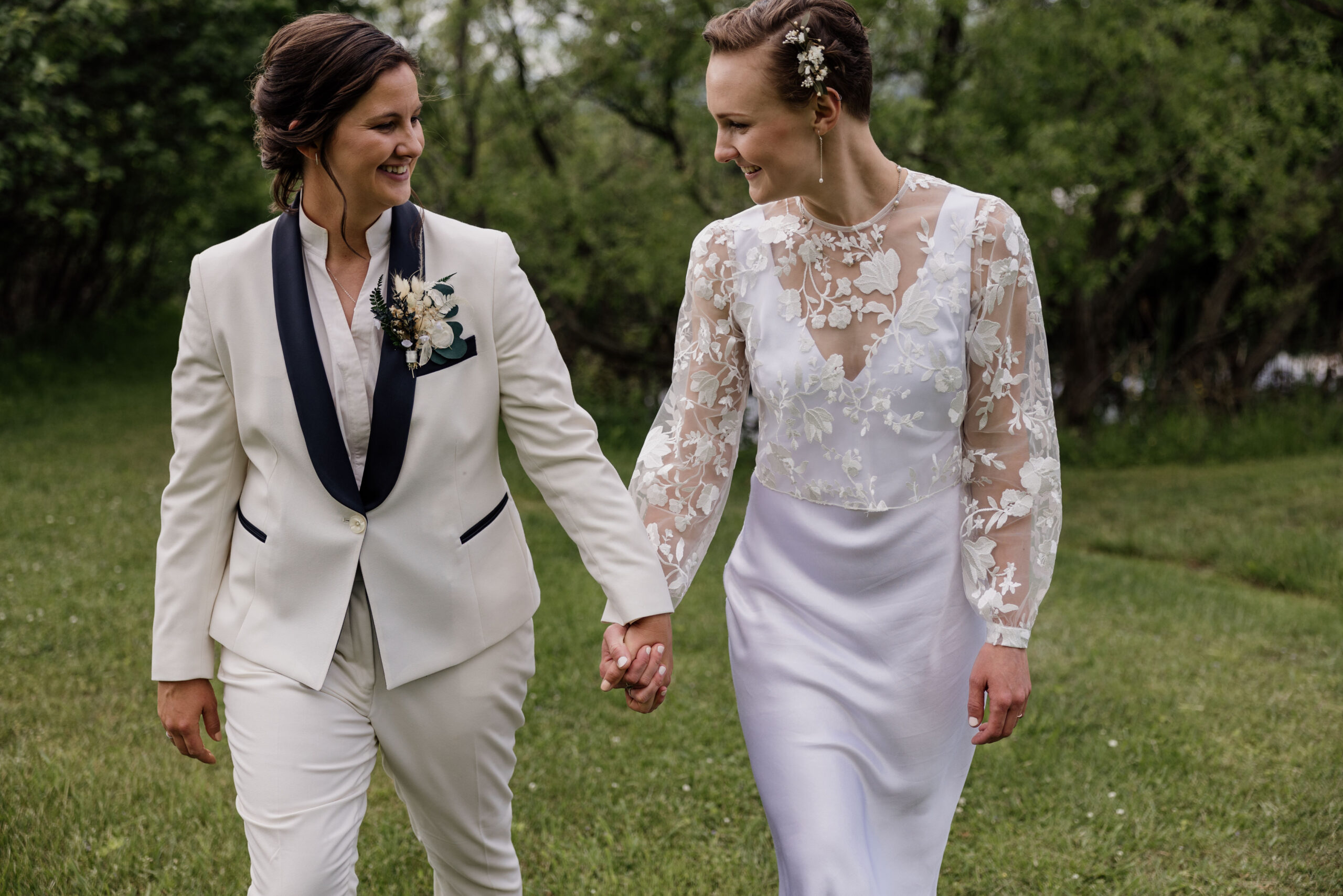 A newlywed couple walking hand-in-hand on a grassy field. One bride is wearing a white suit jacket with black lapels, adorned with a boutonniere, while the other is in a white satin dress with floral lace detailing on the bodice and sleeves. Both are smiling warmly at each other, surrounded by lush greenery in the background.