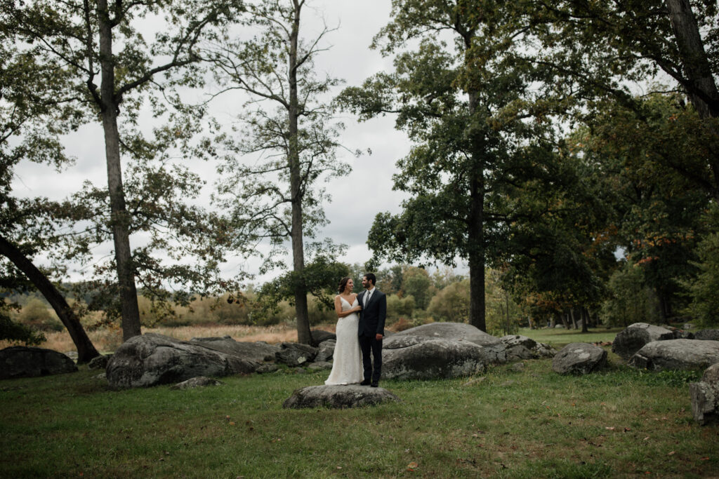 The same couple standing together in the middle of an open, grassy area surrounded by trees. The bride is wearing a white wedding dress, and the groom is in a dark suit. They gaze at each other lovingly, with large boulders and tall trees framing the scene.