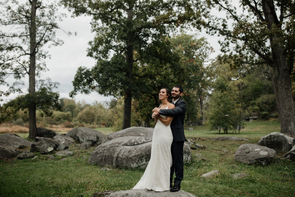 A bride and groom standing together on a large rock in a forested park. The bride is wearing a white, sleeveless wedding dress, and the groom is dressed in a dark suit. He stands behind her, holding her in an embrace, while they both look into the distance, surrounded by lush greenery and large rocks.