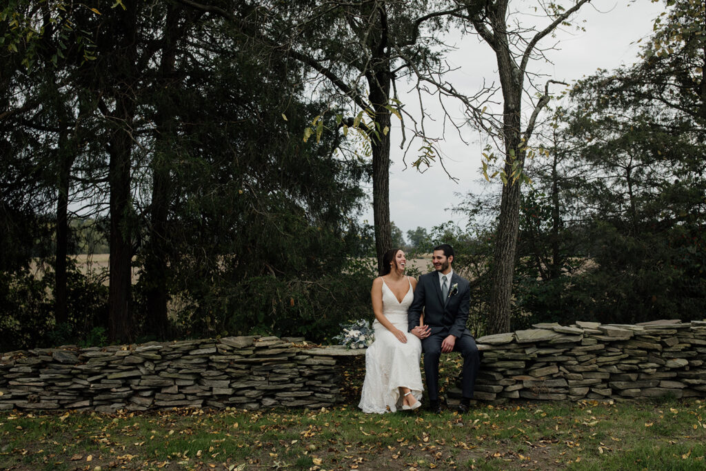 A bride and groom sitting on a stone wall while holding hands. The bride is wearing a sleeveless dress, and the groom is wearing a suit and tie. 