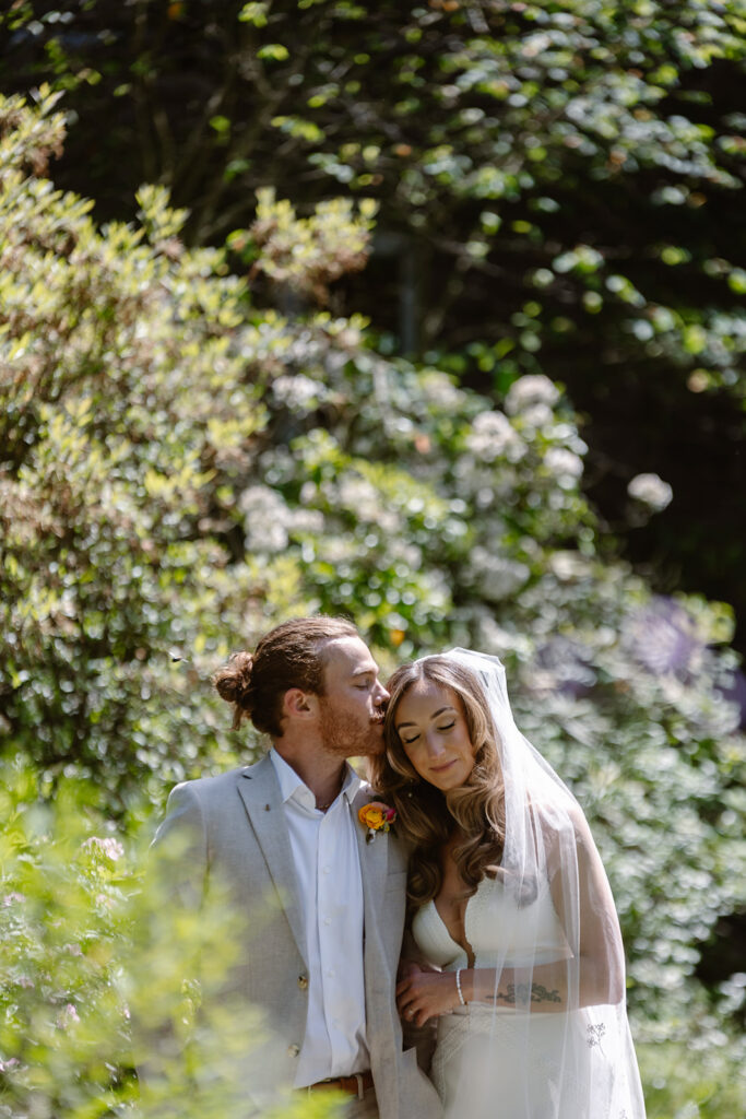 The bride and groom take a quiet moment in a lush flower garden during their wedding.  The brides eyes are closed, and the groom is gently kissing her forehead. 