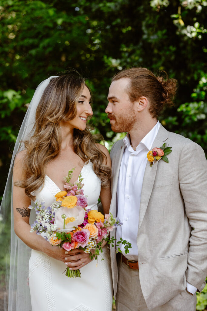 A bride and groom looking at each other on their wedding day. The bride is wearing a stunning Boho dress, and the groom is wearing a tan linen suit. The bride is holding a bright and bold bouquet with large poppies and ranunculus flowers