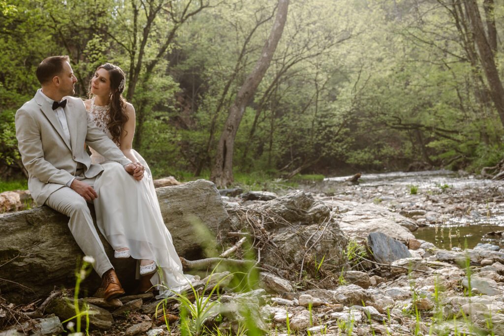 A bride and groom sitting on a large rock by a tranquil river. The bride, in a lace dress, leans towards the groom, who wears a light-colored suit. They are surrounded by a serene, green forest, creating a peaceful atmosphere.