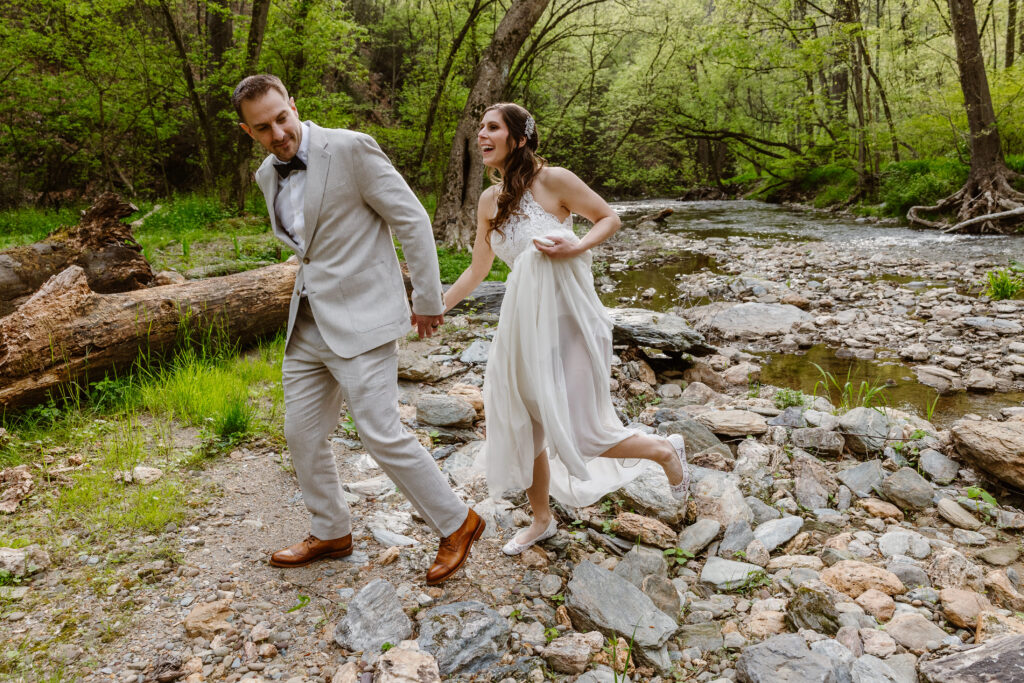 A bride and groom walking hand-in-hand along a rocky riverbank. The groom wears a light suit, and the bride is in a flowing white dress. They are smiling, with the bride playfully lifting her dress as they navigate the rocky terrain.