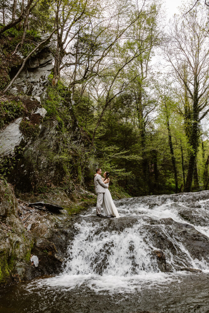 A bride and groom standing together on a rocky waterfall. The groom is in a light suit, and the bride wears a lace wedding dress. They embrace in a romantic moment, surrounded by cascading water and lush greenery.