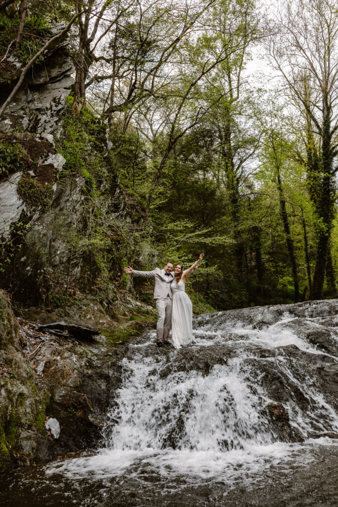 A bride and groom standing together on a waterfall, with water cascading around them. The groom is dressed in a light suit, while the bride wears a flowing white dress. They look joyful, raising their hands in celebration amid the natural beauty.