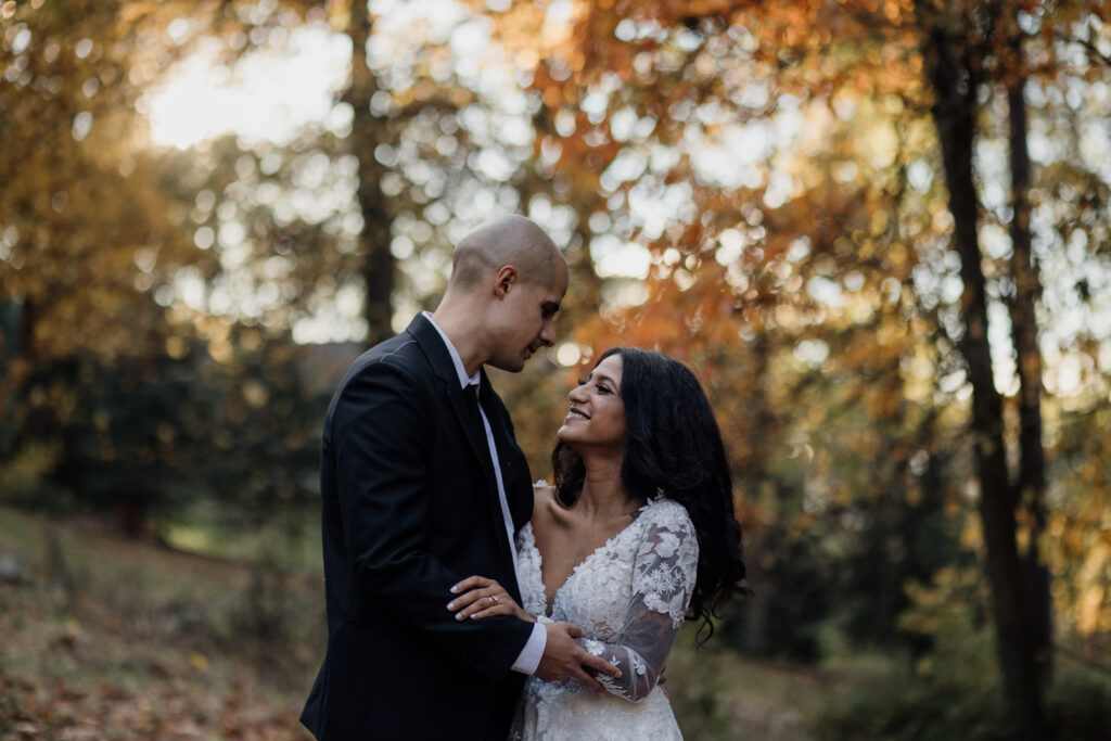 A couple standing in a forested area during the fall, with leaves in warm shades of orange and yellow. The bride is wearing a white lace dress, and the groom is dressed in a black suit. They gaze into each other’s eyes with warm smiles