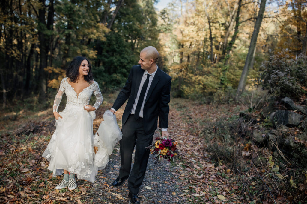 A bride and groom walking hand-in-hand along a forest trail. The bride is wearing a lace wedding dress with sneakers, and the groom is dressed in a black suit. He holds a vibrant bouquet with red and orange flowers as they walk together.