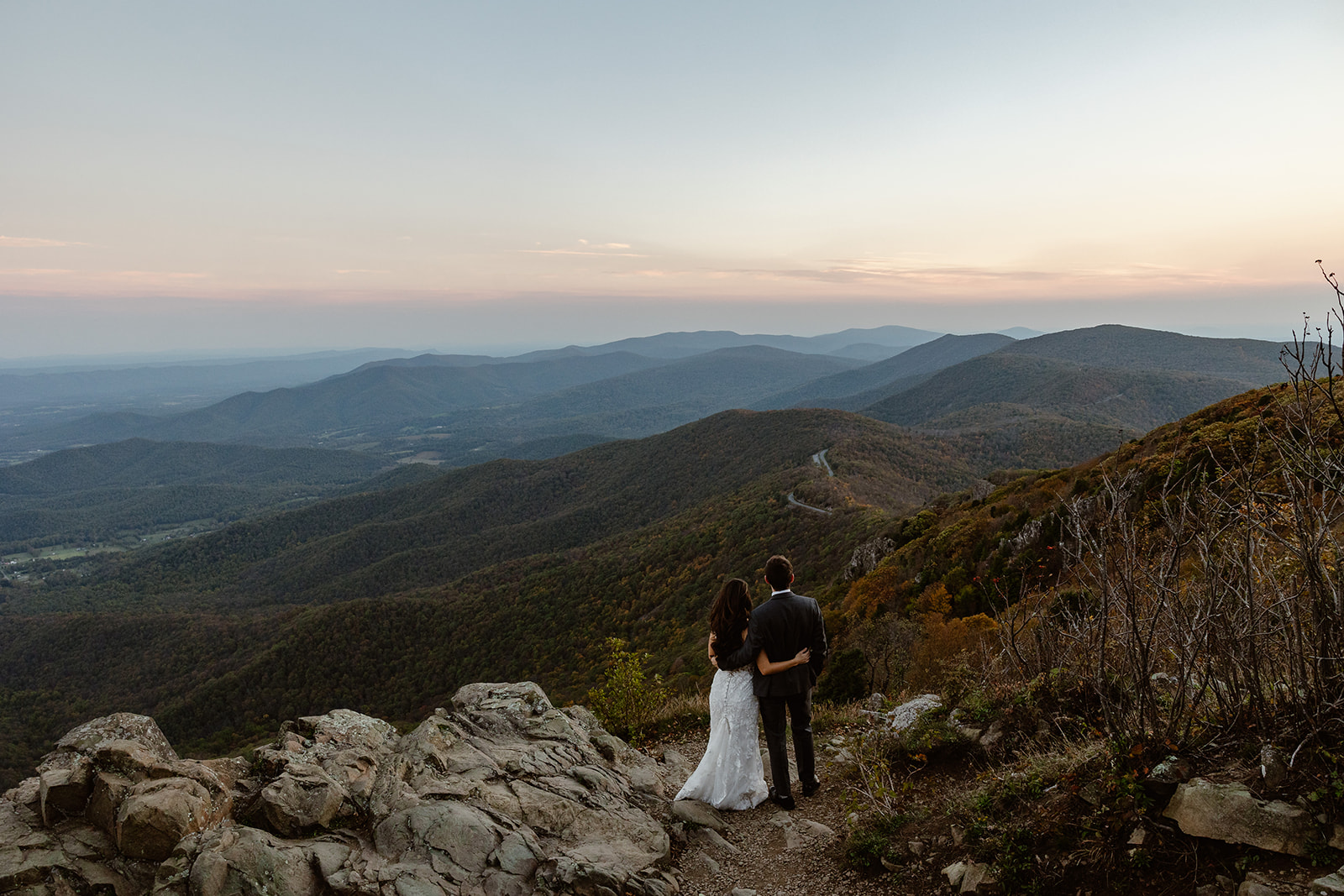 A couple stands with their arms around each other, taking in the view at Stony Man trail in Shenandoah. It is sunrise, and it looks like the leaves are starting to change. She is wearing a delicate lace dress, and he is in a black suit.