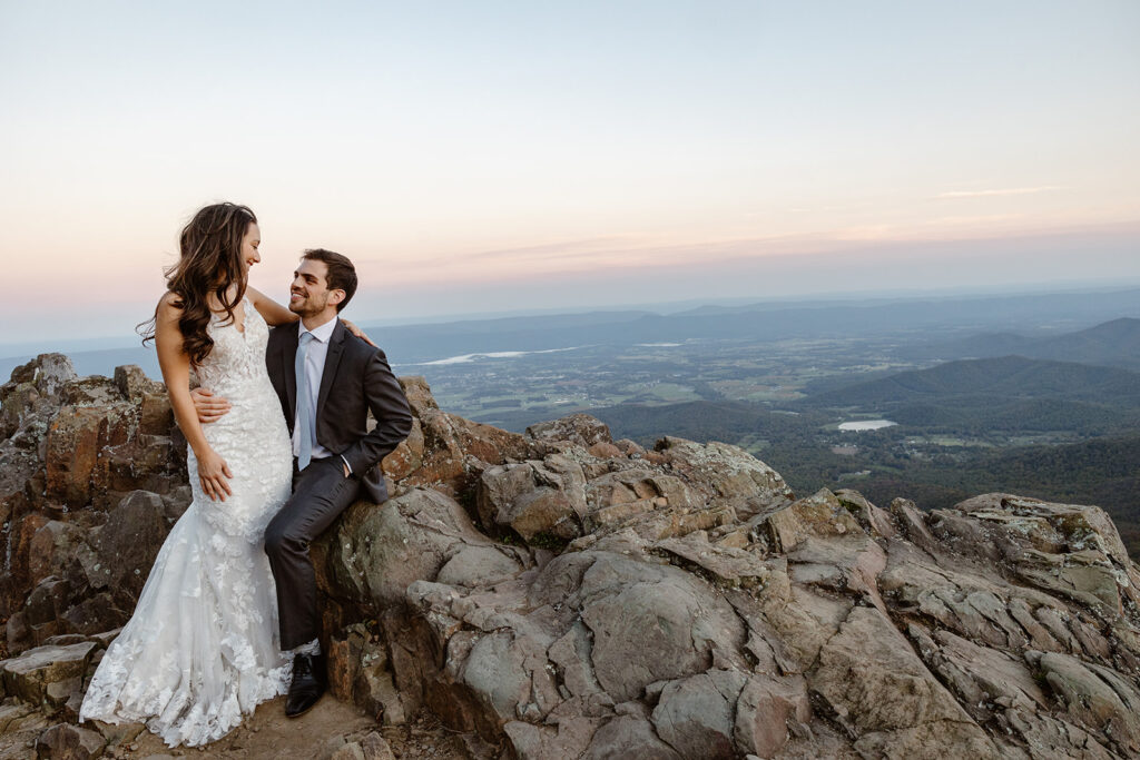 An eloping couple sitting on some rocks at Stony Man Overlook. She is sitting on the groom's lap, and they are exchanging sweet smiles. It is blue hour, and the sky is full of hues of blue and pink.