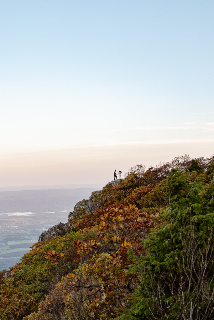 A couple is standing on the top of a rocky mountain. The groom is holding the bride's hand trying to help guide her to the edge safely. The photos is taken from a far distance away so the couple looks tiny amongst the vast mountainous backdrop