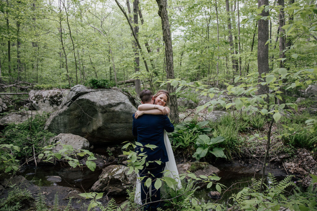 A bride and groom embrace in a forest setting, surrounded by dense green foliage, rocks, and a small creek. The bride is smiling warmly over the groom’s shoulder as they hold each other close. This image conveys a sense of intimacy and the beauty of a natural, serene environment.