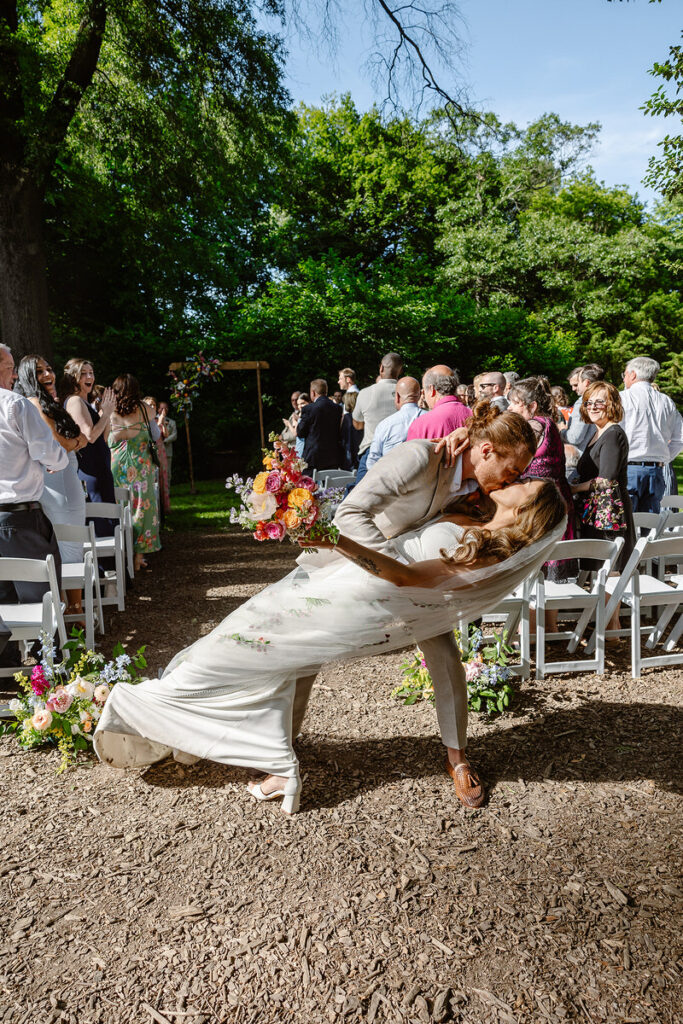 The bride and groom share their vows under a floral-adorned wooden arch in an outdoor wedding ceremony. The tall trees and vibrant green leaves create a magical canopy, while the gathered guests look on, seated in white chairs, witnessing this special moment. The bright sunlight filters through, giving the scene a warm, inviting glow.