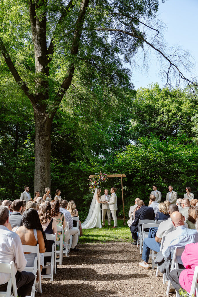 A couple stands beneath a white portico at the Awbury Arboretum. The bride is wearing a flowing gown with delicate floral embroidery, while the groom is dressed in a light-colored suit. The lush greenery and tall trees frame the background, capturing the serene and romantic ambiance of the moment.