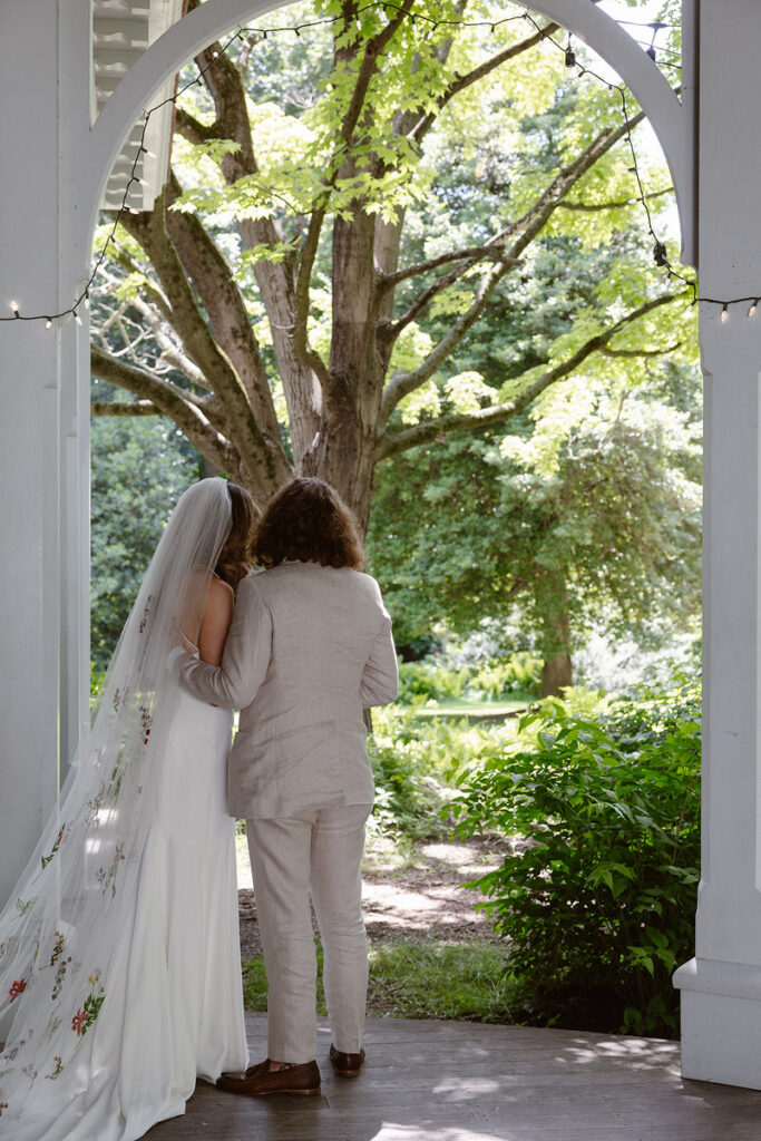 The couple is captured from behind, standing closely together under the arch of the portico. The bride's long veil with embroidered flowers cascades down her back. Soft sunlight filters through the leaves of a large tree, adding a dreamy feel to this intimate scene.