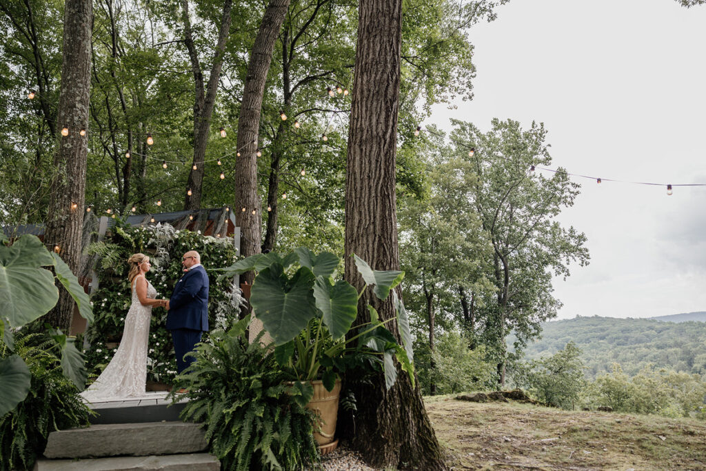 A couple standing together on a rustic outdoor platform surrounded by lush greenery and string lights, exchanging vows. The bride is wearing a lace gown, while the groom is dressed in a navy suit, with a backdrop of trees and hills in the distance.