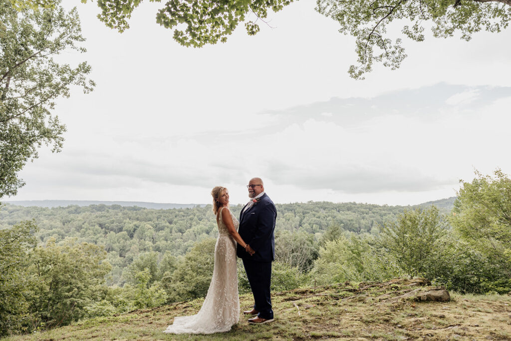 The bride and groom hold hands and smile, standing on a scenic hilltop with a panoramic view of forested hills under an overcast sky. The bride wears a lace dress, and the groom is dressed in a dark suit.