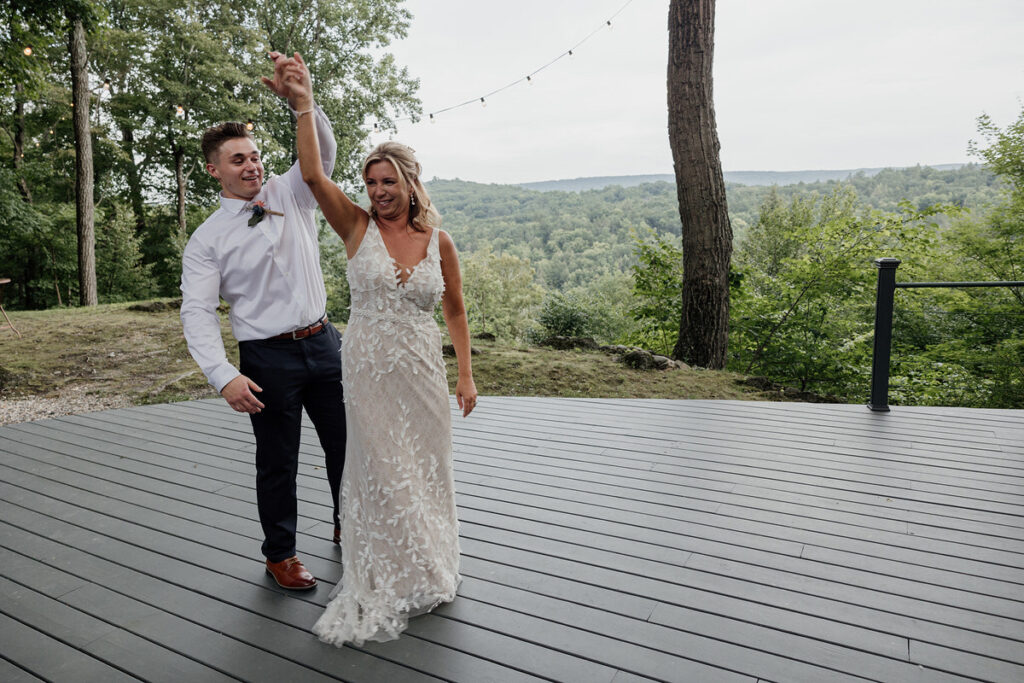 The bride dances with her son, both smiling and holding hands, on a wooden platform with a forested view in the background. The bride is in a floral lace gown, and the guest is wearing a white shirt and dark trousers.