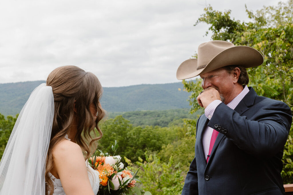 A bride stands facing her father, who is wearing a cowboy hat and suit, in an emotional moment before the wedding. She holds a bouquet of peach and white flowers, while the father looks touched, covering his mouth with his hand. The scenic mountain landscape is visible in the background.