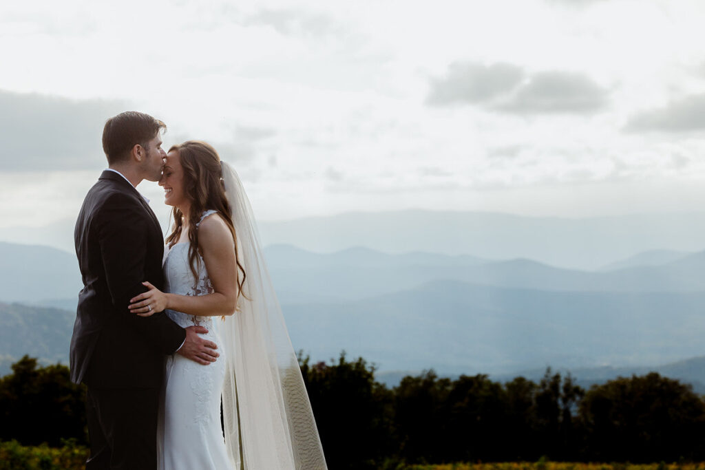 The bride and groom share an intimate moment, with the groom kissing the bride’s forehead against a backdrop of mountains and clouds. They hold each other close, smiling and looking deeply content.