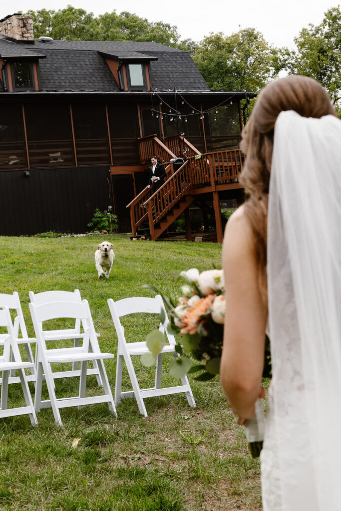 A bride stands with her back to the camera, holding a bouquet as a dog runs towards her on a grassy lawn. In the background, a groom stands on a wooden staircase in front of a dark house, looking down at the scene. White folding chairs are set up for an outdoor ceremony.