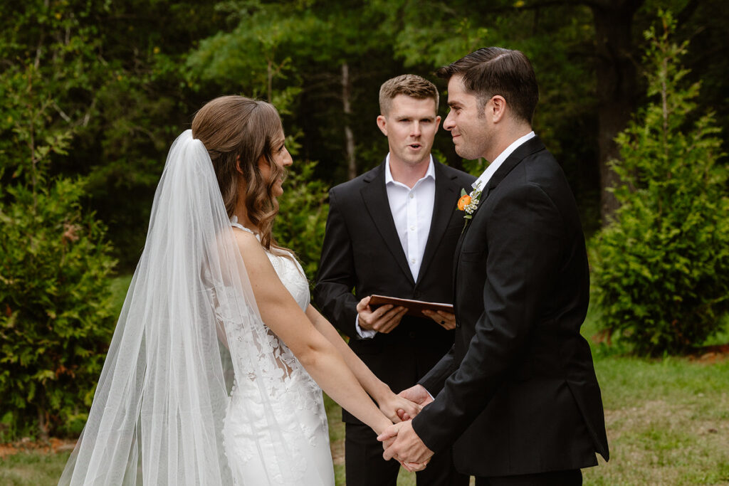 The bride and groom hold hands and face each other, exchanging vows in an outdoor setting. The groom wears a black suit with a boutonniere, and the bride is in a white dress with a long veil. An officiant stands in the background, holding a book, surrounded by greenery.