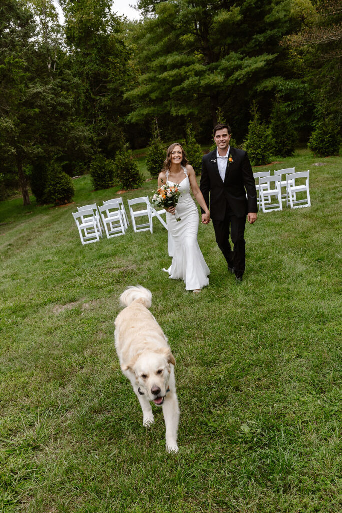 A bride and groom walking hand-in-hand down the aisle, smiling happily. The bride is holding a bouquet of peach and white flowers, while a golden retriever walks in front of them. White chairs are arranged in the background on a grassy outdoor setting.