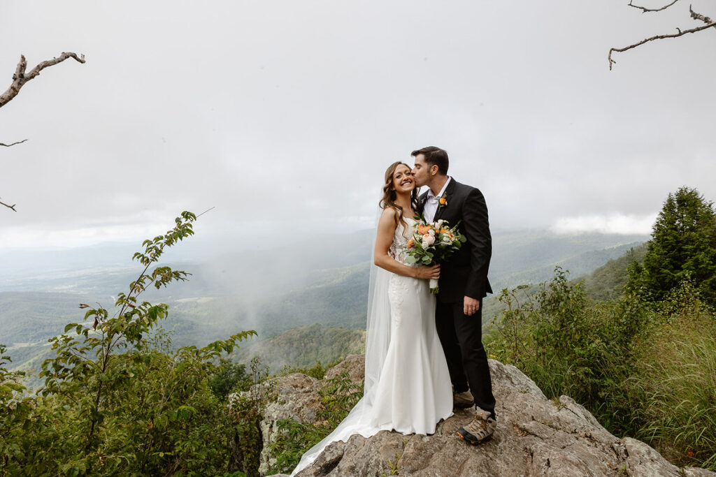 The bride and groom share a kiss on top of a rocky cliff with a scenic, cloud-covered mountain view in the background. The bride holds a bouquet of peach and white flowers, and they both look joyful amidst the stunning landscape.