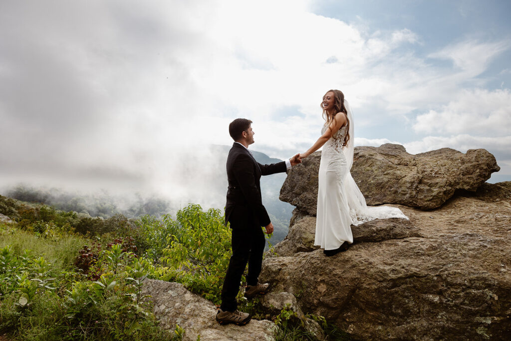 The bride stands atop a large rock, smiling and holding hands with the groom, who stands below her. They are surrounded by lush greenery, with clouds and mountains visible in the background, giving a dreamy and adventurous feel.