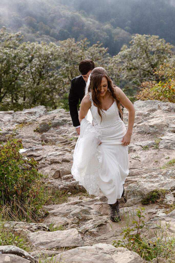 The bride and groom walk hand-in-hand up a rocky path, surrounded by greenery and foggy mountain views. The bride holds up her dress as she climbs, smiling, while the groom follows closely behind.